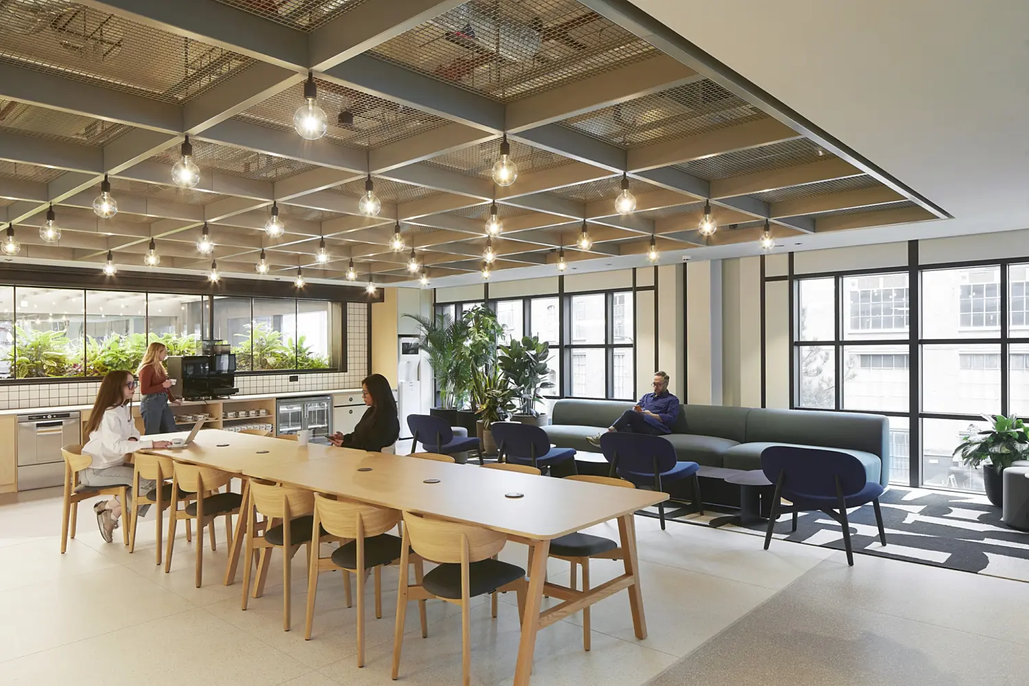 Spacious open plan office breakout area with a long wooden communal table and chairs in the foreground, a sofa seating area with blue chairs and green bench seating to the right, and a kitchenette and planting along the back wall, lit by a grid ceiling with exposed structure and hanging lights, with several people seated or standing throughout the space and large windows along one side.