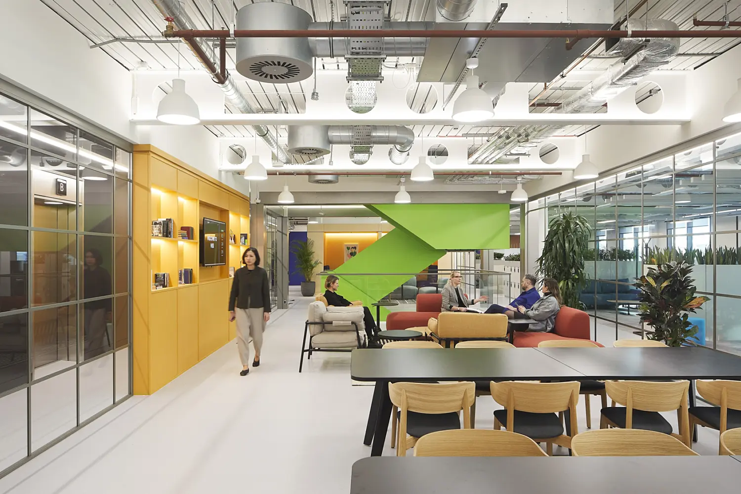 Open plan office breakout and dining area with a long black table and wooden chairs in the foreground, a yellow shelving wall and glass meeting rooms on the left, and a central lounge with sofas arranged around low tables in front of a green staircase, with exposed ceiling services, pendant lighting, indoor planting and several people seated and walking through the space.