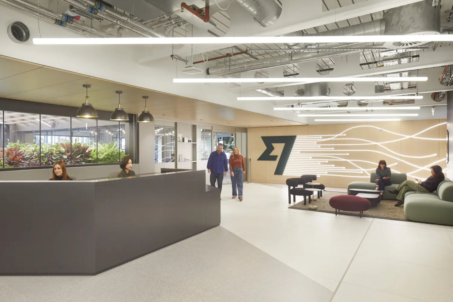 Contemporary office reception and breakout area with a long dark reception desk on the left, exposed ceiling services and linear lighting above, and a seating area on the right with sofas and low tables, backed by a feature wall with illuminated graphic lines, while several people are seated or walking through the space and a planted area is visible behind glass at the rear.