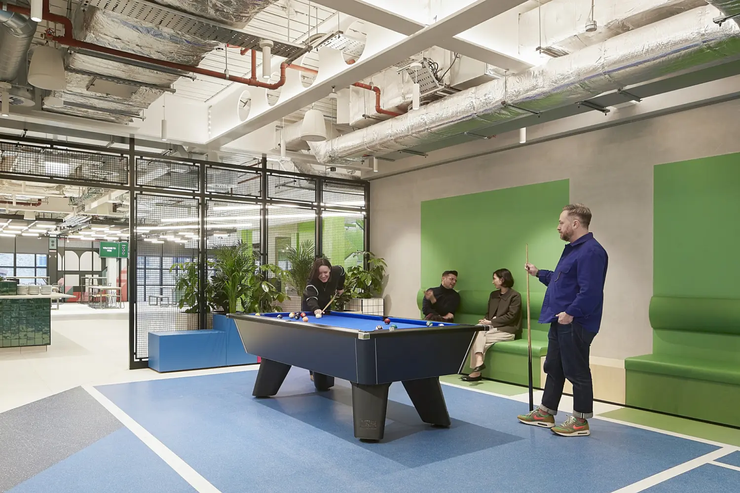Office games and breakout area with a blue pool table set on a marked blue floor, green upholstered bench seating along the wall where several people are sitting, and a person standing beside the table, all beneath a ceiling with exposed ductwork, pipes and lighting, with glass partitions and indoor planting visible in the background.