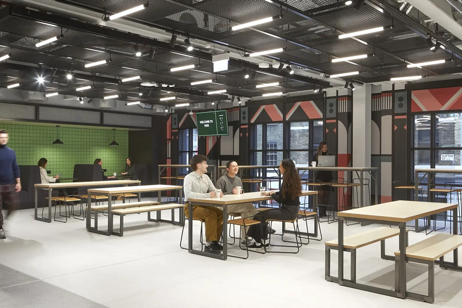 Open plan office breakout area with long communal tables and bench seating arranged across a light floor, a green tiled wall with additional seating on the left, patterned feature walls and large windows on the right, and an exposed ceiling with grid lighting, with several people seated at tables and others walking through the space.
