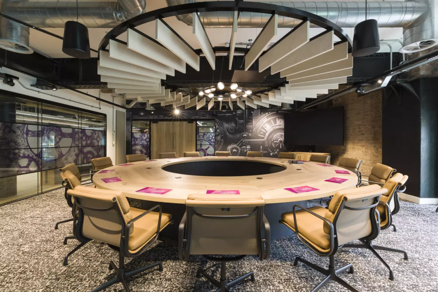 Modern meeting room with an industrial design featuring exposed ductwork and brick walls. A large circular conference table with a central opening dominates the space, surrounded by tan leather swivel chairs. Above the table, a striking ceiling installation of angled acoustic panels radiates outward from a black circular frame, paired with a contemporary chandelier. The flooring is patterned carpet in neutral tones, and the walls include glass partitions with purple graphic designs and a black feature wall with technical illustrations. The overall look combines bold geometry with functional acoustic elements.