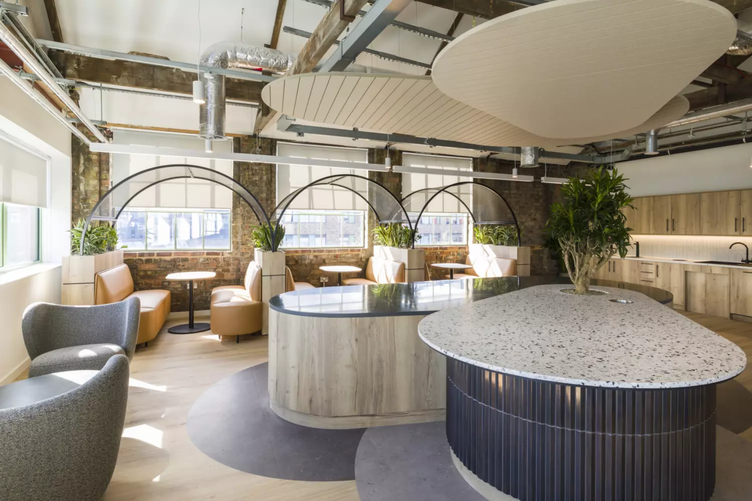 Contemporary office breakout area with an industrial design featuring exposed brick walls, visible steel beams and ductwork. The space includes curved wooden booths with tan leather seating arranged under large arched black metal frames, complemented by round tables. A central island with a terrazzo countertop and ribbed dark base adds texture, while a second island with a light wood finish integrates greenery. Overhead, acoustic panels in soft beige tones contrast with the raw ceiling structure. Natural light streams through wide windows with roller blinds, creating a bright and inviting atmosphere.