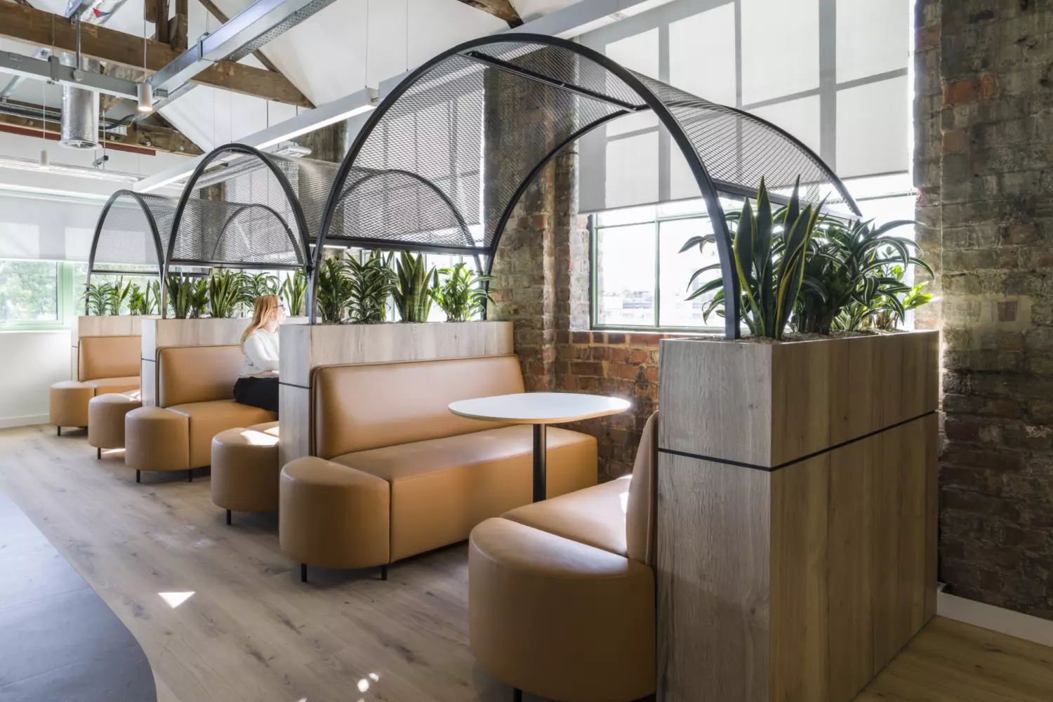 Office breakout space with an industrial design featuring exposed brick walls, steel beams and visible ductwork. The area includes curved wooden booths with tan leather seating arranged under black metal arches, paired with round tables. Two large islands dominate the centre: one with a terrazzo countertop and ribbed dark base, the other in light wood with integrated greenery. Overhead, acoustic panels in soft beige tones contrast with the raw ceiling structure. Natural light streams through wide windows with roller blinds, creating a bright and welcoming atmosphere.