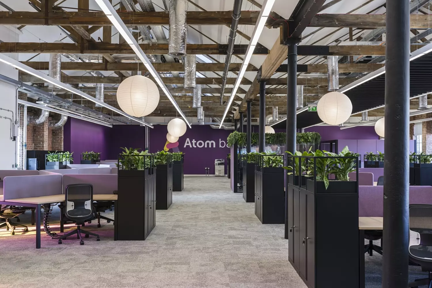 Open-plan office with an industrial aesthetic featuring exposed timber beams, visible ductwork and suspended linear lighting. The space is divided into workstations with purple upholstered panels and black ergonomic chairs. Rows of black planter cabinets with lush greenery create natural partitions. The flooring is soft grey carpet, and the walls are painted in a deep purple tone with the text “Atom bank” visible in the background. Large round paper lanterns hang from the ceiling, adding a warm contrast to the raw structural elements. The overall design blends natural textures with vibrant colour for a collaborative workspace.