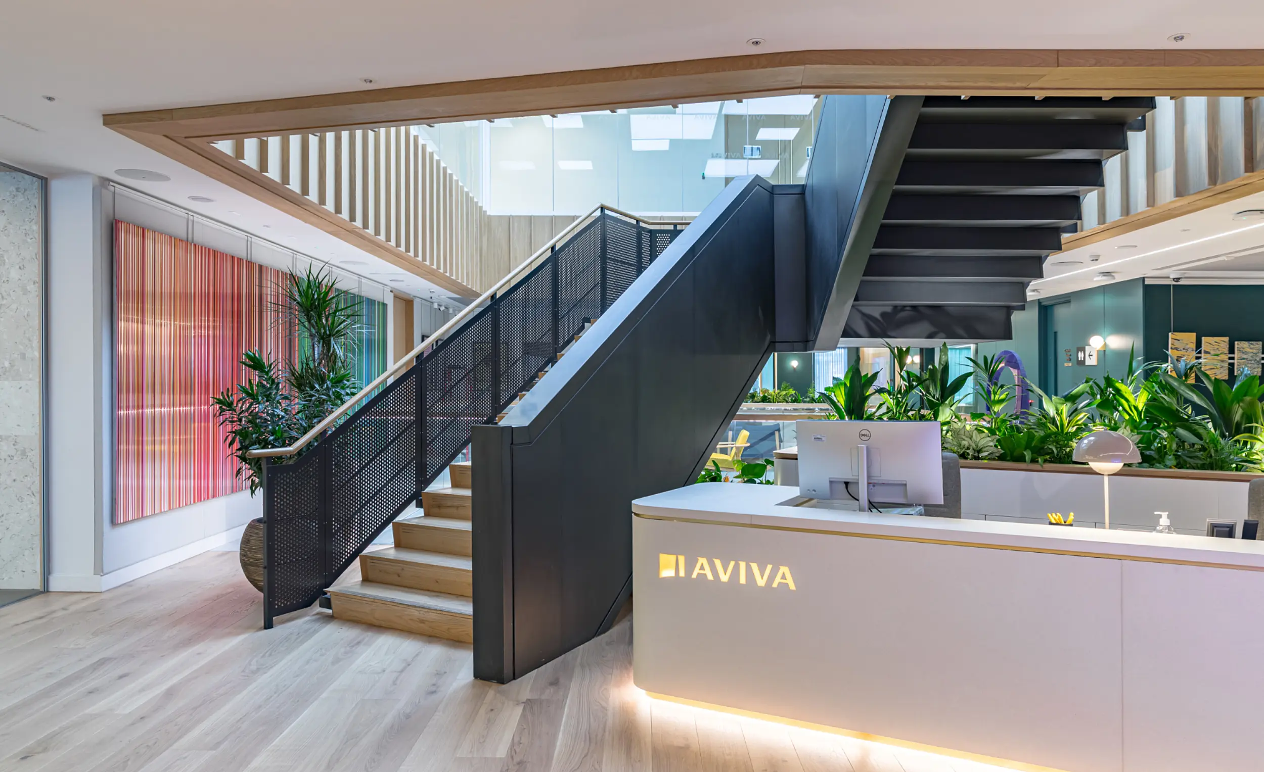 A contemporary reception area with light wood flooring and a curved white desk displaying the Aviva logo in illuminated lettering. Behind the desk is a row of lush green plants forming a natural divider. A wide staircase with wooden steps and black metal balustrades rises to an upper level, framed by a timber‑lined opening in the ceiling. The space is brightly lit by natural light from above and features modern architectural details, including clean lines, glass panels and colourful vertical artwork on the left wall.