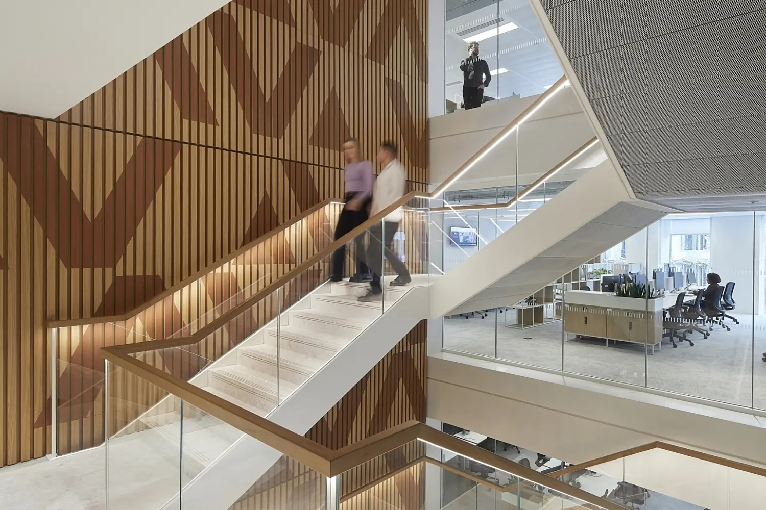 A multi level office interior with an open staircase featuring glass balustrades and light stone steps. The staircase runs beside a tall wall covered in vertical timber slats arranged in a geometric pattern. Several people are walking on the stairs and others are visible in the open plan workspace on the right, which has desks, task chairs and large windows. The space is bright with a mix of wood and glass finishes.