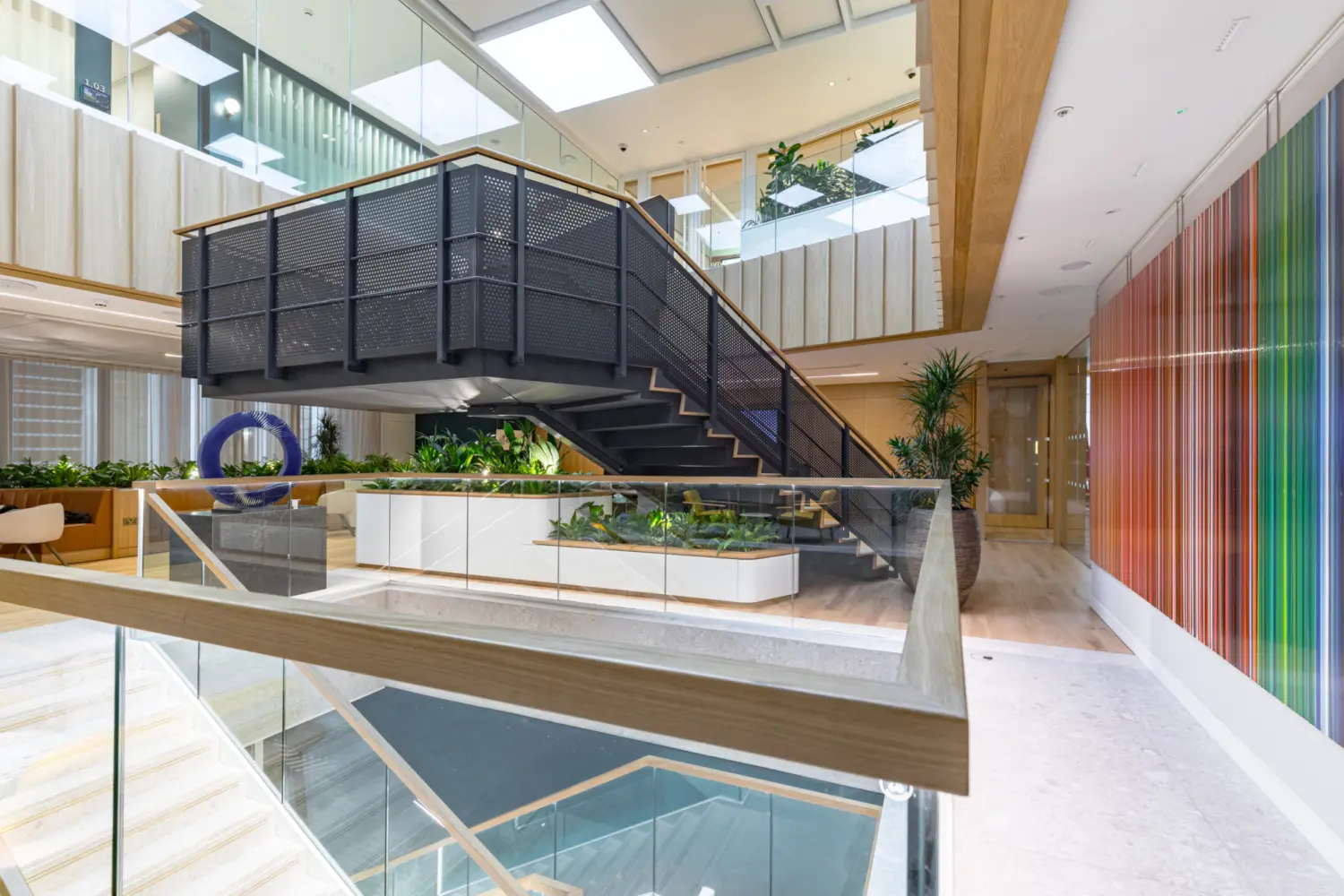 A bright multi level office atrium with a central black metal staircase and glass balustrades. The space features light wood finishes, large planters with greenery and an open mezzanine above. To the right is a tall wall of vertical coloured panels. Natural light enters from overhead skylights, and there are work areas and seating zones visible around the atrium.