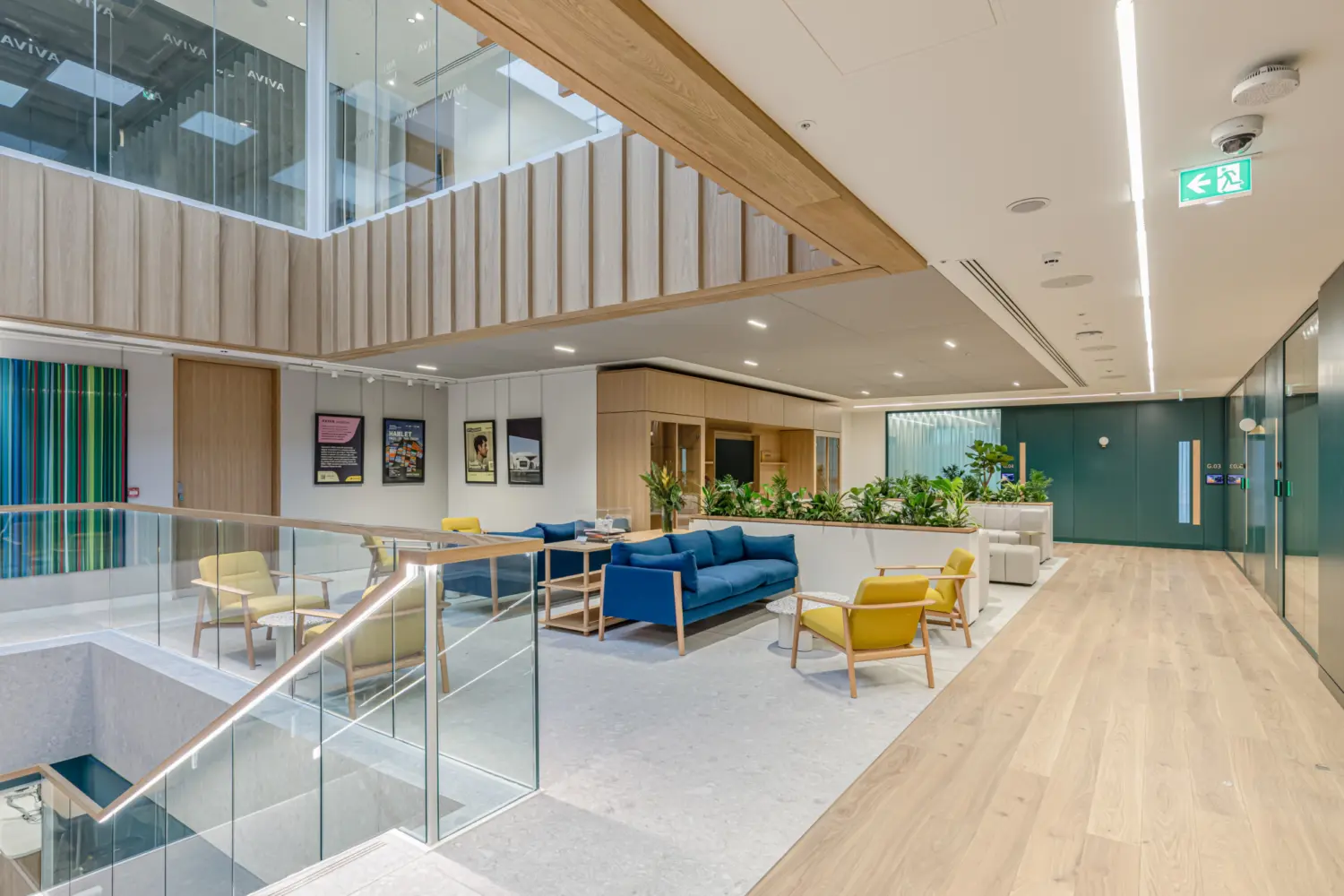 A bright open landing area in an office with light wood flooring and a glass balustrade overlooking a stairwell. The space is furnished with blue and yellow lounge chairs, a blue sofa and small tables arranged in a casual seating zone. Behind the seating are planters filled with greenery and a wall with framed artwork. To the right, a corridor with teal doors leads to other rooms. Above, a large opening reveals the floor above with wood slat detailing and glass panels. The overall setting is modern and spacious.