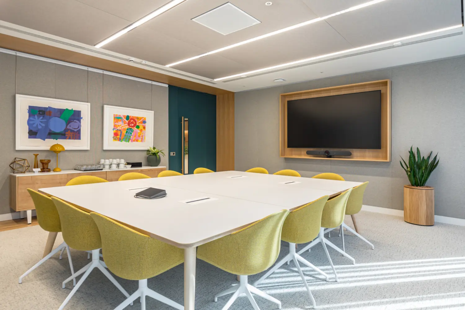 A bright meeting room with two white tables arranged together and surrounded by yellow swivel chairs. A large wall mounted screen with a wooden frame is positioned at the front of the room. On the left wall are two colourful framed artworks above a sideboard that holds drinks, cups and decorative items. A tall plant sits in the corner on the right. The room features soft ceiling lighting, a neutral colour scheme and a teal door at the back.