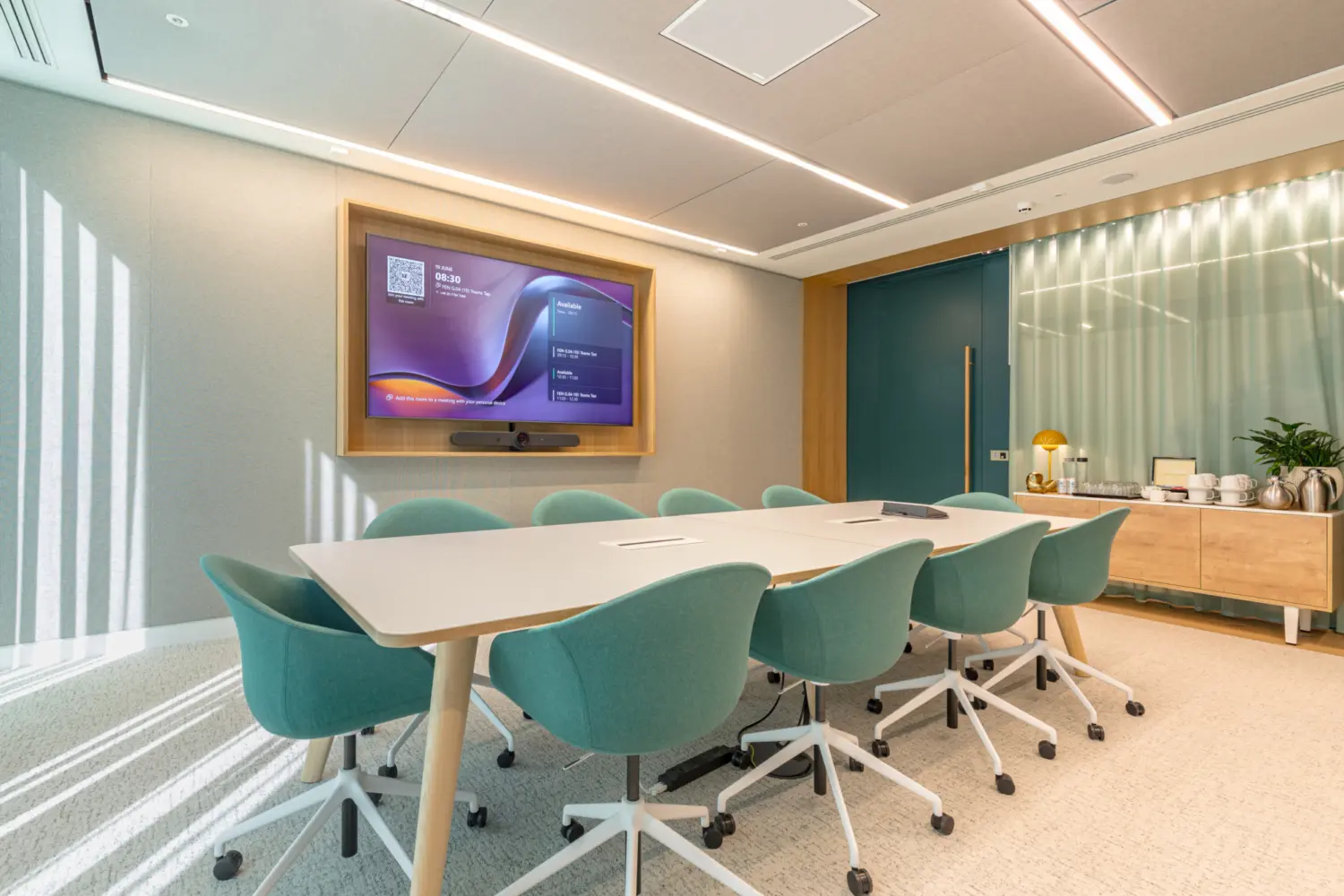 A small modern meeting room with a long table surrounded by teal swivel chairs. A large wall mounted screen at the front displays a digital meeting interface. Soft ceiling lighting and a clean neutral colour scheme give the room a bright feel. To the right is a sideboard with drinks, cups and a lamp, set against a frosted glass partition and a teal door.