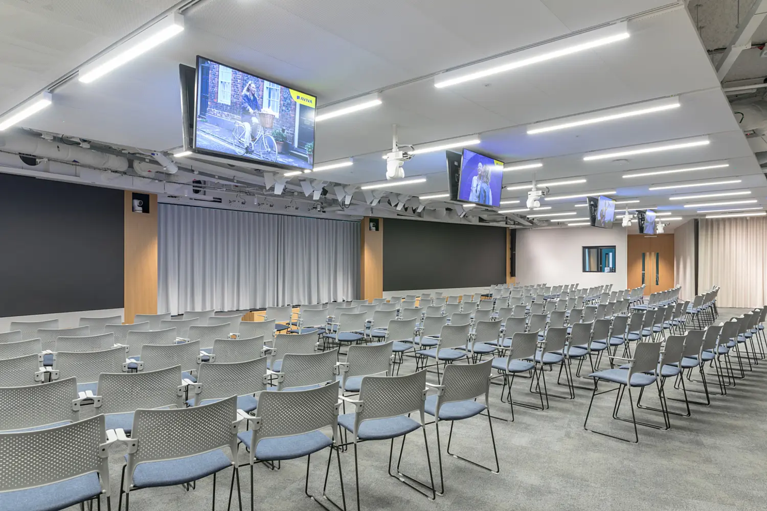 A large conference room arranged with rows of grey chairs facing a stage area with wide black presentation screens and closed curtains. Multiple ceiling mounted monitors hang above the seating, displaying video content. The space has bright even lighting, exposed ceiling details and a neutral colour palette. The room appears set up for a talk or presentation.