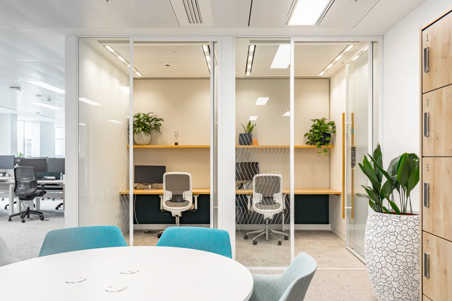 A small enclosed workspace with two individual desk pods separated by glass partitions. Each desk has a task chair, a monitor and a wooden shelf with plants placed above. The pods are fronted by full height sliding glass doors with subtle patterned detailing. In the foreground is a round meeting table with teal chairs. To the right stands a tall white planter with a leafy plant, and lockers line the adjacent wall. The wider open plan office is visible through the glass on the left.