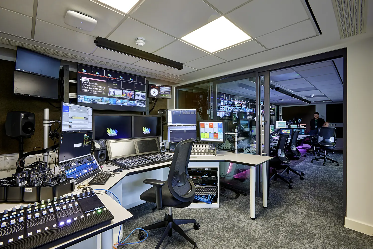 A professional broadcast control room filled with technical equipment and multiple screens. In the foreground, a curved workstation is covered with audio and video control panels, mixing desks and several computer monitors displaying broadcast software and graphics. A black office chair sits in front of the central console, with visible cabling and equipment racks stored beneath the desk.  Above the workstation, a row of mounted screens shows various live feeds, graphics and news content. The ceiling features a suspended lighting fixture and standard office ceiling panels with recessed lights.  To the right, a large glass partition reveals an adjacent control area with additional workstations, monitors and rolling office chairs, where more broadcast equipment is in use. Subtle coloured lighting adds depth to that space. The floor is carpeted in a dark tone, complementing the neutral walls and desks.  The overall setting conveys a busy, high-tech environment designed for live television production, with a focus on monitoring, editing and controlling multiple video and audio sources in real time.