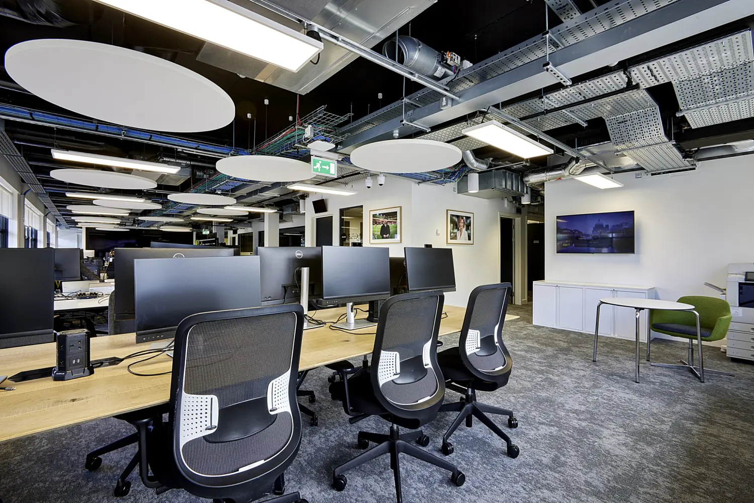 A modern open-plan office workspace with an industrial-style ceiling and a clean, functional layout. In the foreground, a long light wood desk runs horizontally, fitted with several large black computer monitors and paired with black mesh-backed swivel chairs on castor wheels. Power modules and cables are neatly integrated into the desk surface.  Above, the exposed ceiling reveals metal ductwork, cable trays and structural elements, complemented by rectangular light panels and several large white circular acoustic panels suspended at varying heights. The lighting is bright and even, highlighting the technical, contemporary feel of the space.  In the background, additional desks and monitors continue into the open area, suggesting a shared working environment. On the right, a small breakout corner includes a round table with two chairs, one in a muted green tone. Nearby, low white storage cabinets sit against the wall, and framed photographs or artwork hang above them. A printer is positioned at the far right edge.  The floor is covered in a dark grey carpet, contrasting with the lighter wood desks and white walls. A green emergency exit sign is visible overhead in the distance. The overall design combines practical workstation layouts with exposed services and acoustic features to create a bright, collaborative office environment.