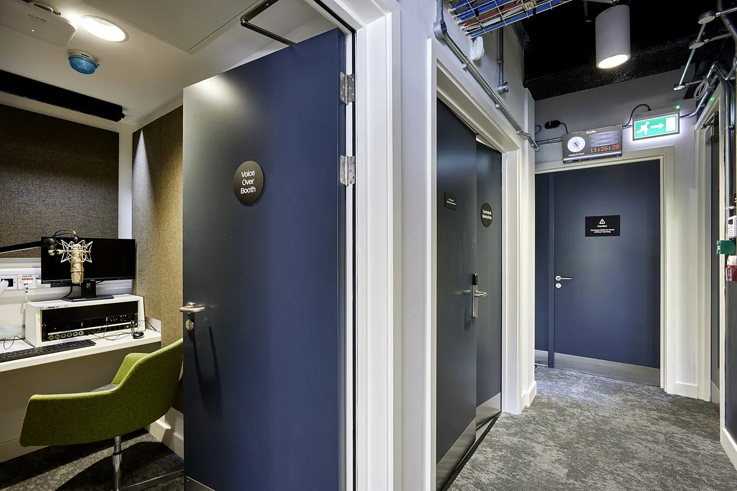 A compact recording and production corridor within a broadcast or media facility. On the left, an open door labelled “Voice Over Booth” reveals a small recording room fitted with acoustic wall panels, a desktop computer, audio equipment and a mounted studio microphone on an adjustable arm. A green upholstered chair is positioned at the workstation.  In the centre and right, a narrow corridor leads past several dark blue doors with signage, each likely marking separate recording or edit rooms. Above one of the doors, a wall-mounted digital clock and timer display is visible, alongside a green illuminated emergency exit sign.  The ceiling features exposed cable trays, ducts and suspended lighting fixtures, while the walls are finished in a mix of white and dark panels. The floor is covered in a dark carpet, helping to absorb sound. The overall space feels functional and technical, designed for audio recording, editing and broadcast production within a controlled environment.