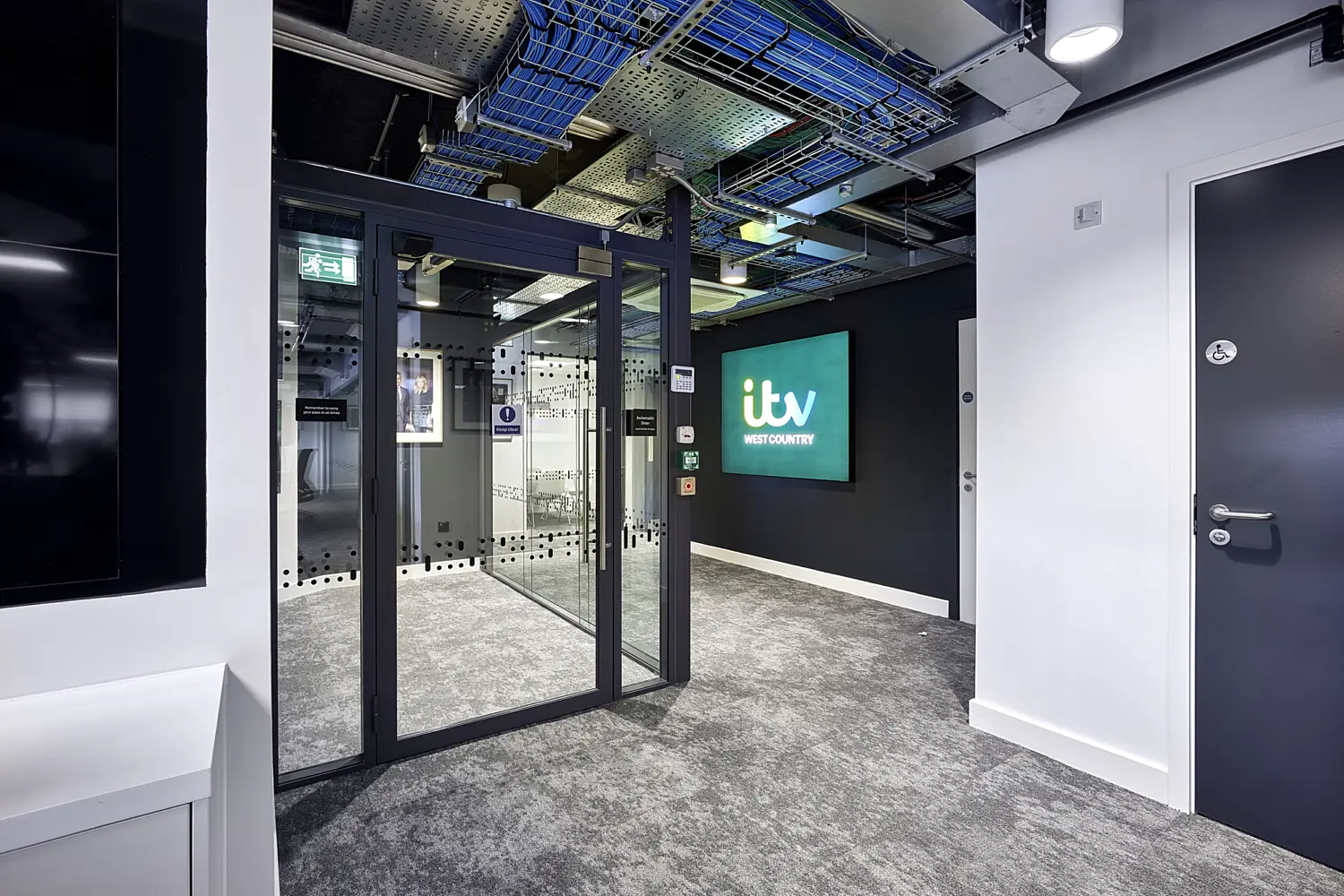 A modern office reception area with a contemporary, technical design. In the centre, a set of full-height glass double doors with dark metal frames leads into another space, with a dotted privacy pattern applied across the glass panels. Beyond the doors, additional glazed partitions and a framed wall image are visible.  On the right, a dark wall features a large screen displaying the text “itv WEST COUNTRY” against a green and blue background, acting as a focal branding element. Adjacent to this, a solid grey door with accessibility signage is set into a white wall.  Above, the ceiling is left exposed, revealing cable trays, ductwork and structural elements, with integrated linear and spot lighting providing bright, even illumination. The floor is finished in a grey textured carpet, complementing the monochrome and dark-toned palette.  Overall, the space combines glass, metal and neutral finishes with visible building services to create a clean, professional entrance area suited to a media or broadcast environment.