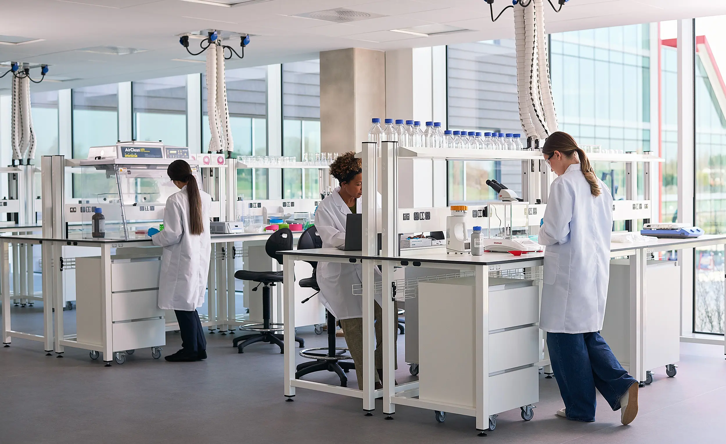 Bright, modern laboratory with large floor-to-ceiling windows, white workstations and scientific equipment. Three researchers in white lab coats work at separate benches, using laboratory instruments and computers. Shelving holds rows of sample bottles and glassware, while ceiling-mounted extraction arms hang above the work areas. The space is clean, open and filled with natural daylight.
