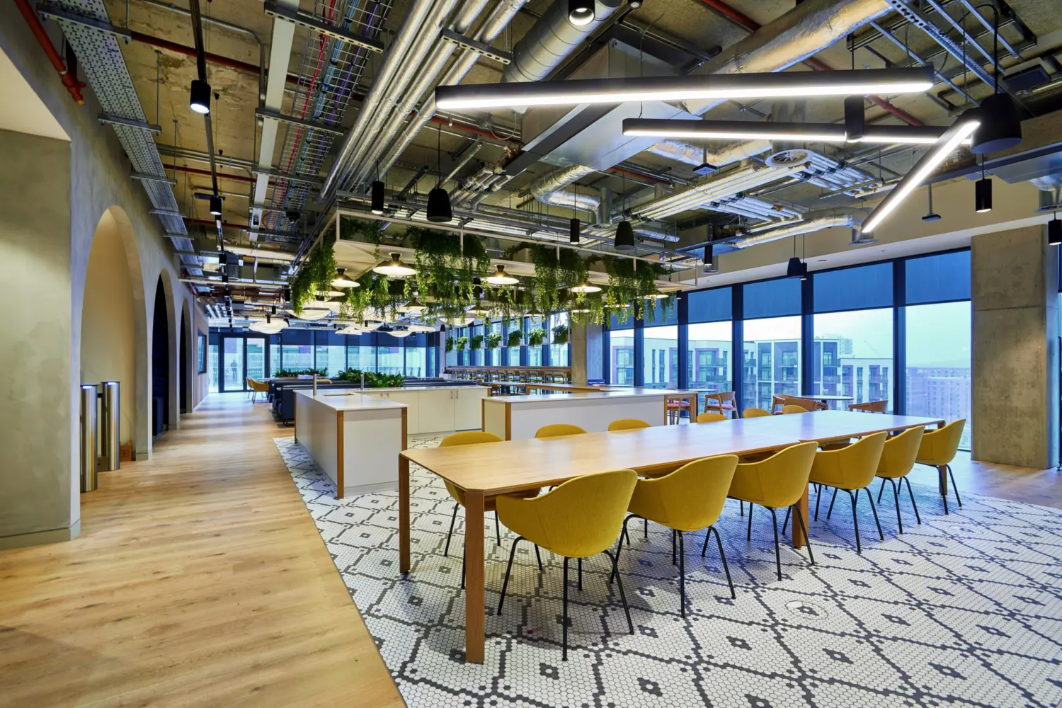Modern open plan office dining area with a long timber table and yellow upholstered chairs set on a patterned tile floor. Exposed ductwork, pipes and linear light fittings cover the ceiling, with hanging plants above a central kitchen island. Large floor to ceiling windows line the right side, while arched concrete openings and light wood flooring run along the left side.
