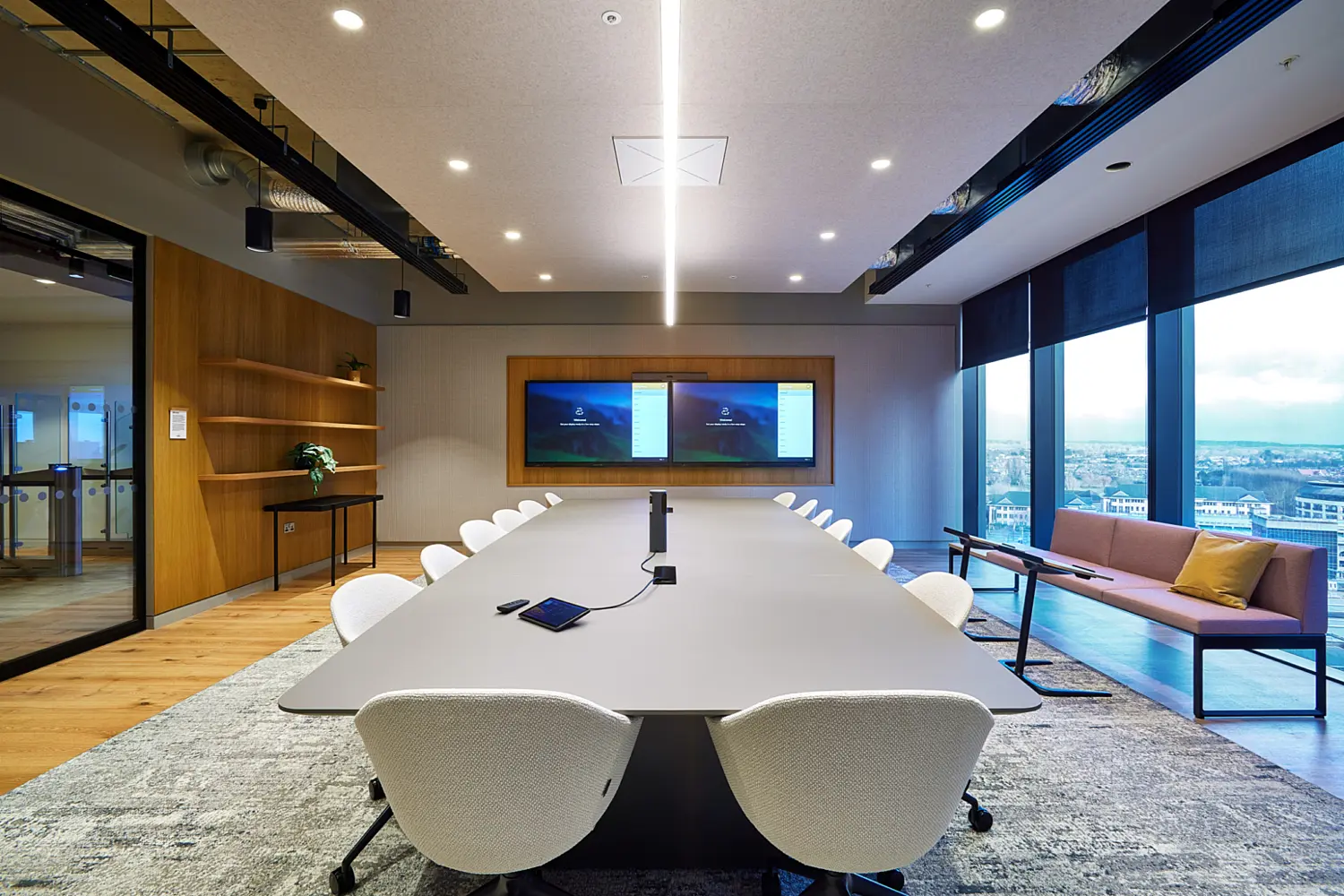 Formal meeting room with a long oval conference table surrounded by white task chairs on a textured rug. Linear ceiling lighting and recessed spotlights provide even illumination, with exposed services along the perimeter. A large wall mounted display sits at the far end, floor to ceiling windows line the right side with city views and a small lounge seat is positioned beside the glazing.