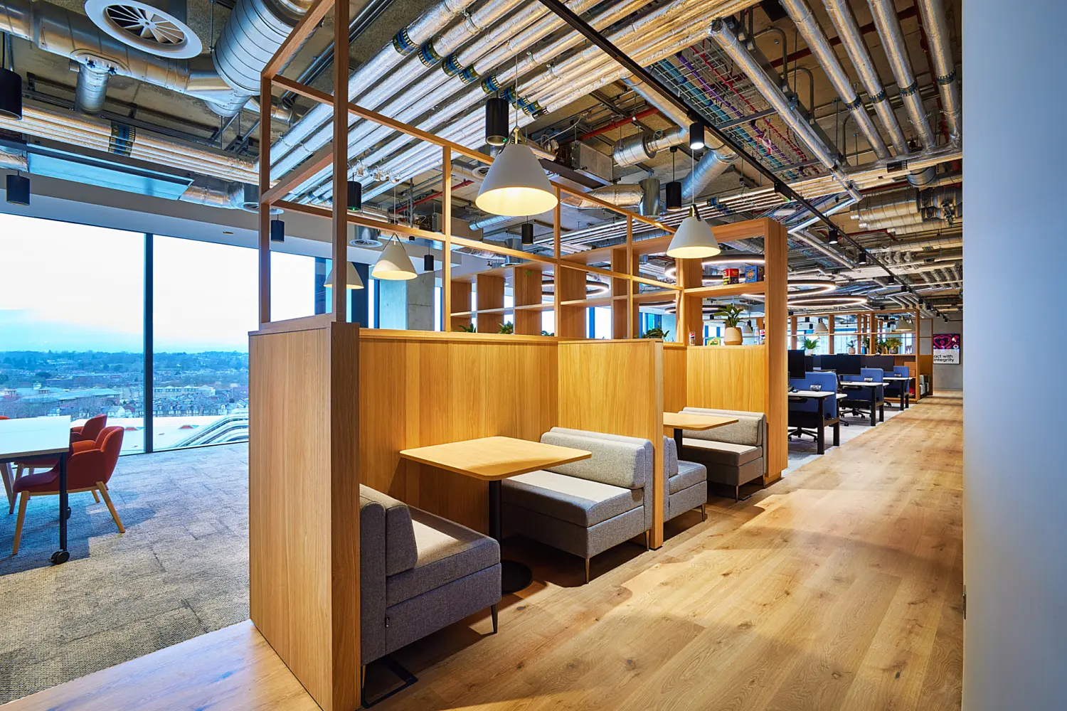 Office breakout booths formed by timber screens with built in seating and small tables arranged along a light wood floor walkway. Grey upholstered benches sit beneath white pendant lights, with exposed ductwork and services overhead. Large windows on the left provide views across the surrounding landscape, while additional flexible seating and work areas extend into the background.