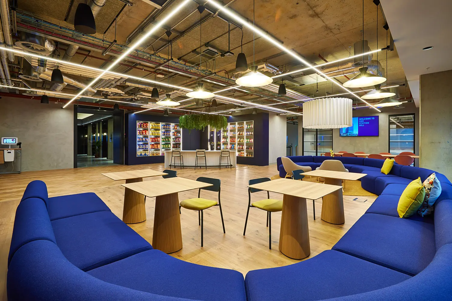 Open plan office lounge with curved blue sofa seating arranged around small timber tables and mixed colour chairs on a light wood floor. Bright linear lighting, pendant lamps and exposed ductwork span the ceiling. A central refreshment bar with shelving and hanging plants sits at the back, with concrete columns, digital screens and open circulation space surrounding the seating area.