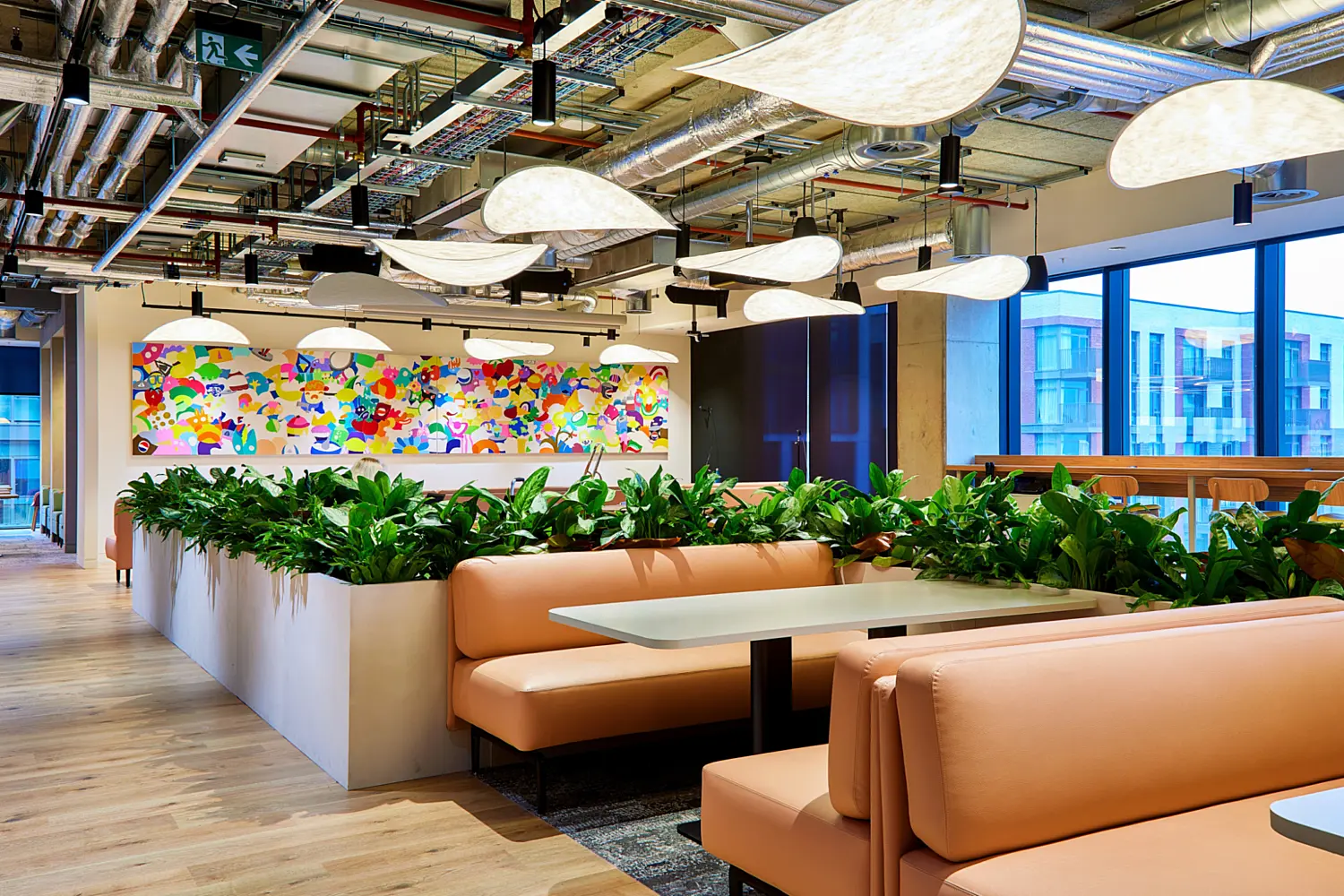 Open plan office lounge and breakout area with soft pink banquette seating around white tables, separated by large planters filled with greenery. Exposed ductwork, pipes and suspended feature lights run across the ceiling. A colourful abstract wall mural sits in the background, with floor to ceiling windows on the right and light wood flooring throughout.