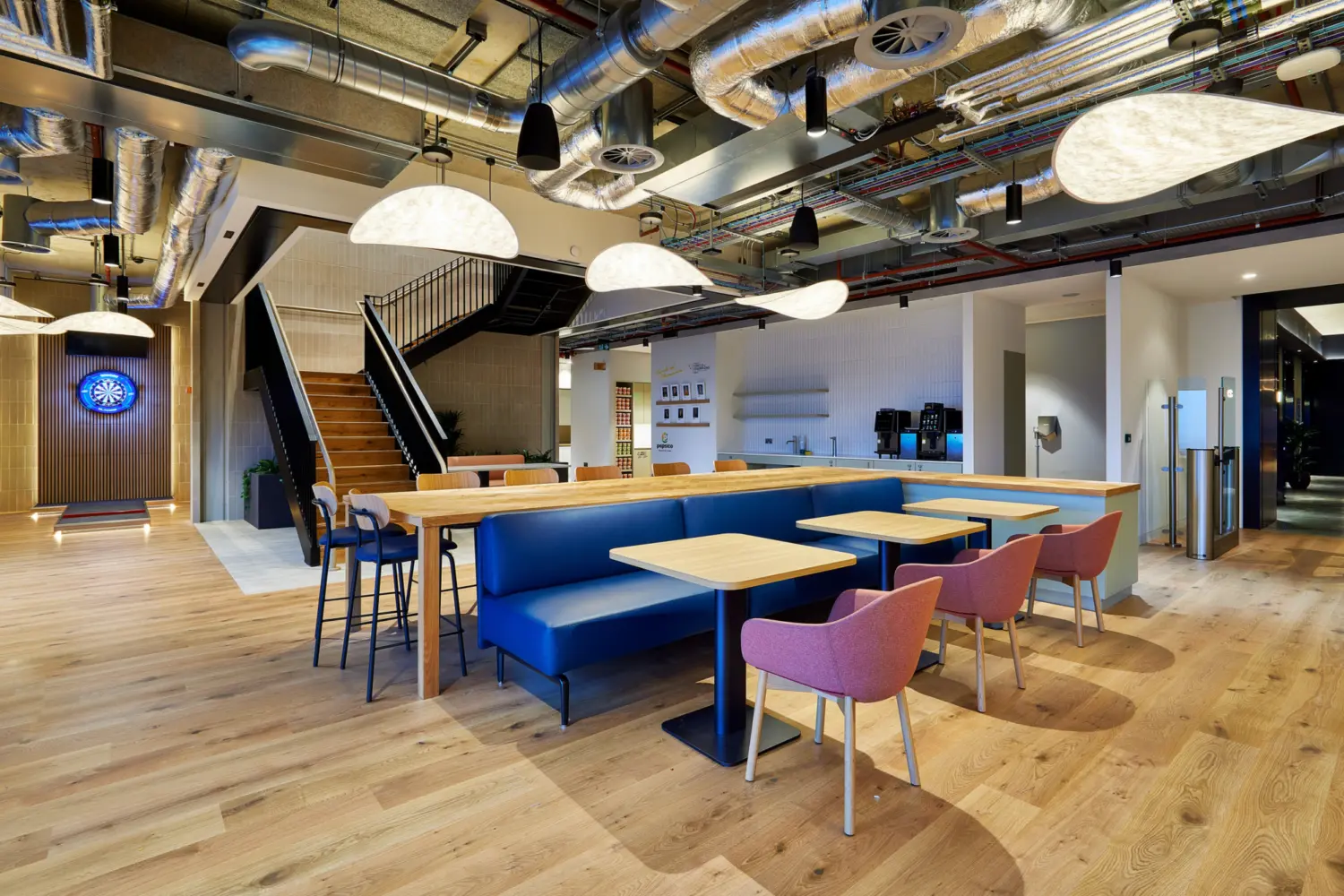 Open plan office breakout area with long communal tables, blue upholstered bench seating and pink chairs on a light wood floor. Exposed ductwork and cables run across the ceiling with large pendant lights. A staircase with black railings leads to an upper level, while a kitchenette with coffee machines sits to the rear.