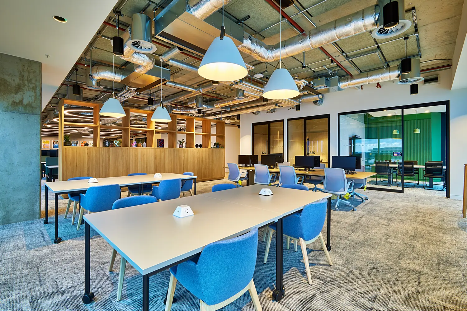 Open plan office work area with shared rectangular tables and blue upholstered chairs set on a grey carpet. Large white pendant lights hang below exposed ductwork and services. Glazed meeting rooms and workstation desks line the right side, with timber joinery and open shelving to the left, creating a bright and functional collaborative workspace.