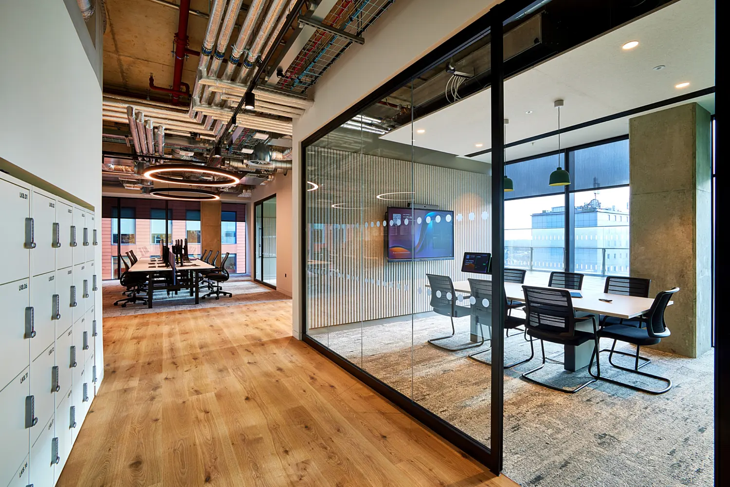 Office corridor with light timber flooring leading past lockers on the left and a fully glazed meeting room on the right. Inside the meeting room is a long table with task chairs set on a textured rug, with circular pendant lighting and a large screen mounted on the wall. Exposed ductwork and services run across the ceiling throughout the space, with large windows at the far end bringing in natural light.