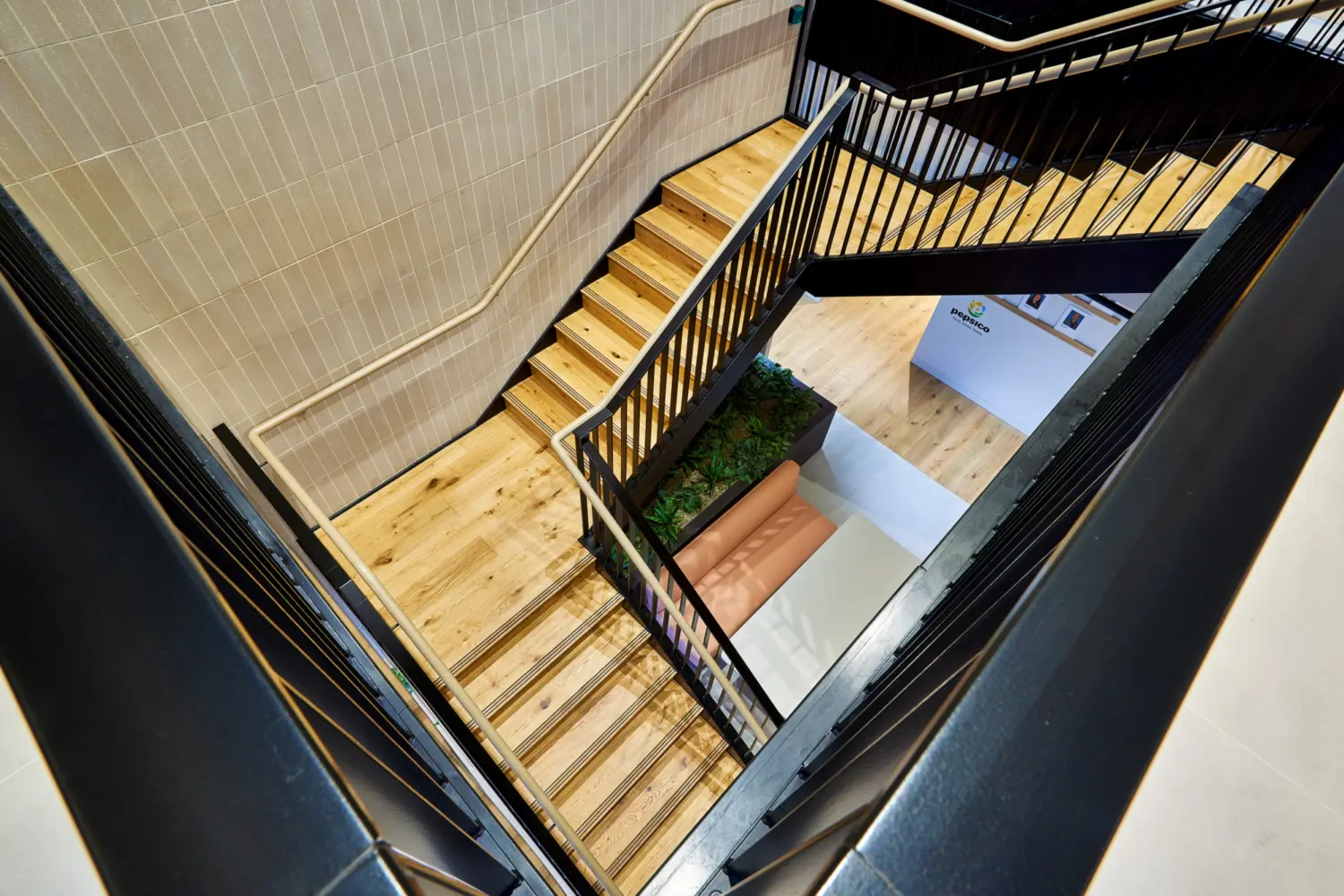 View looking down into an open office stairwell with timber steps and black metal balustrades wrapping around a central void. Light wood flooring continues at each level, with a small seating nook and integrated planter at the base of the stairs. Neutral tiled walls and exposed structure frame the space, creating a bright, vertical circulation area.
