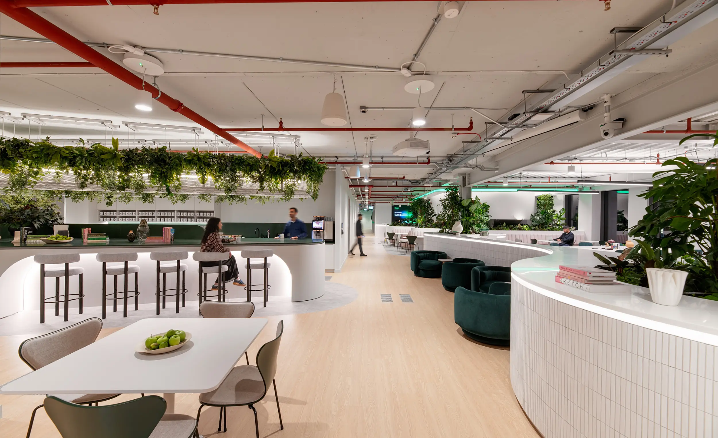 A spacious modern workplace with light wood flooring, white curved counters and abundant greenery. The foreground shows a table with chairs and a bowl of green apples. Further back, a bright bar-style area with high stools sits beneath hanging plants. Several individuals are seated or standing throughout the space. The background includes lounge seating, more plants and illuminated accents that add a soft glow to the open plan setting.