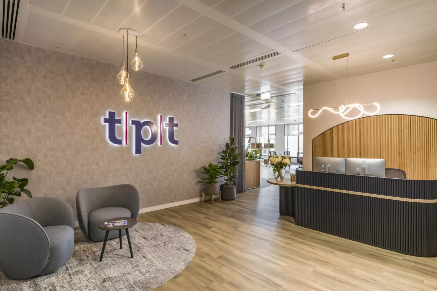 Modern office reception area with wooden flooring, bespoke reception desk, illuminated company logo, grey armchairs on a circular rug, pendant lighting and open plan workspace in the background