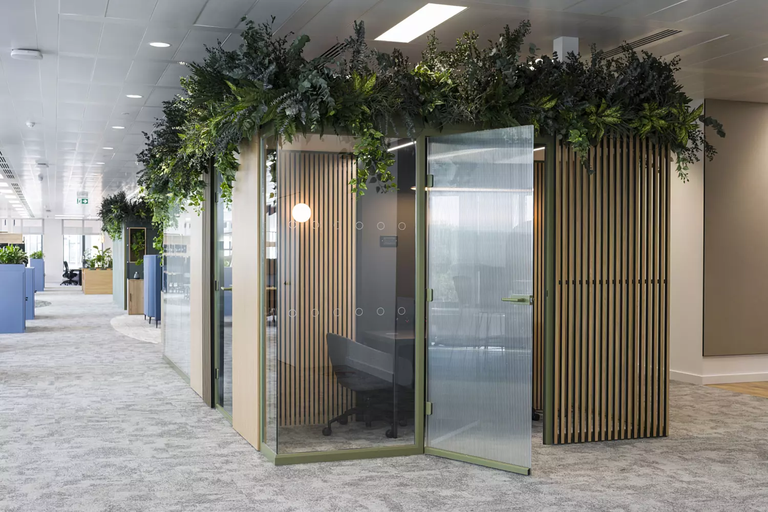 Modern office pod with glass walls, wooden slat detailing, greenery on top and a small table with chair for private work or meetings in an open plan workspace