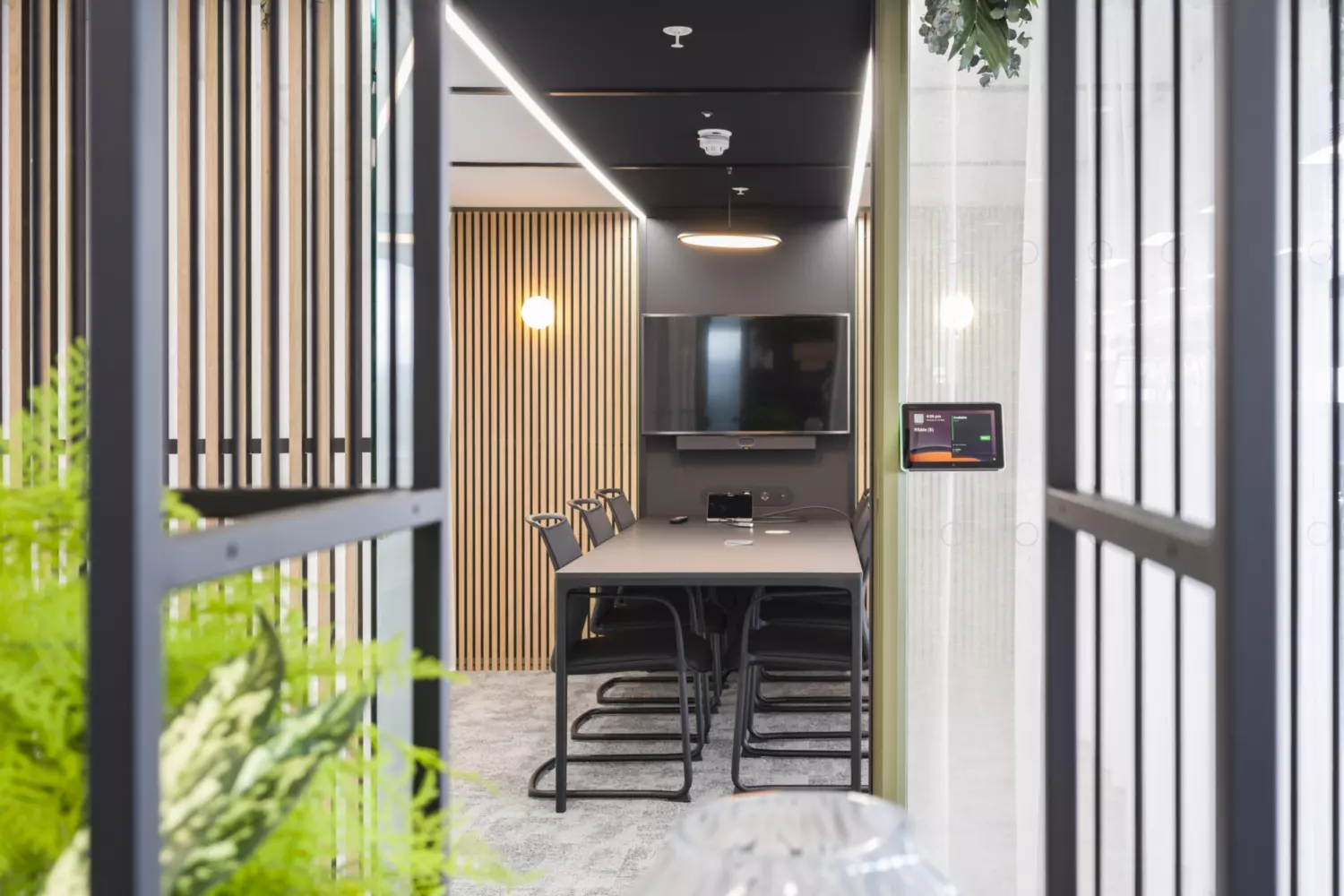 Modern conference room with long meeting table, chairs, wall-mounted screen, wooden slat feature wall, digital booking panel and natural accents for a professional workspace.