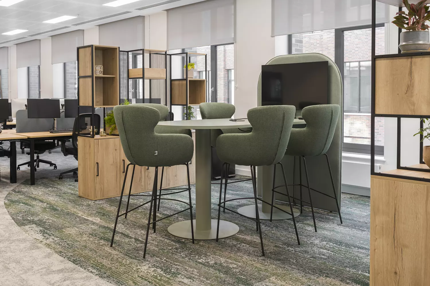 Modern office with round table, green upholstered chairs, semi-private booth seating, patterned carpet, wooden shelving with plants and open-plan desks in a bright hybrid workspace