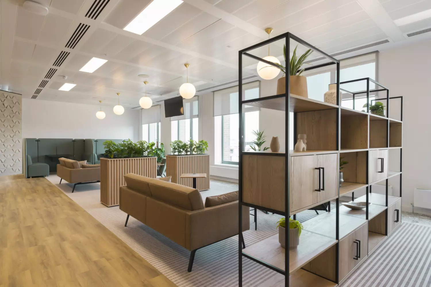 Open plan office lounge with wooden flooring, brown sofas, low wooden tables, decorative shelving with plants and natural light creating a modern collaborative workspace.