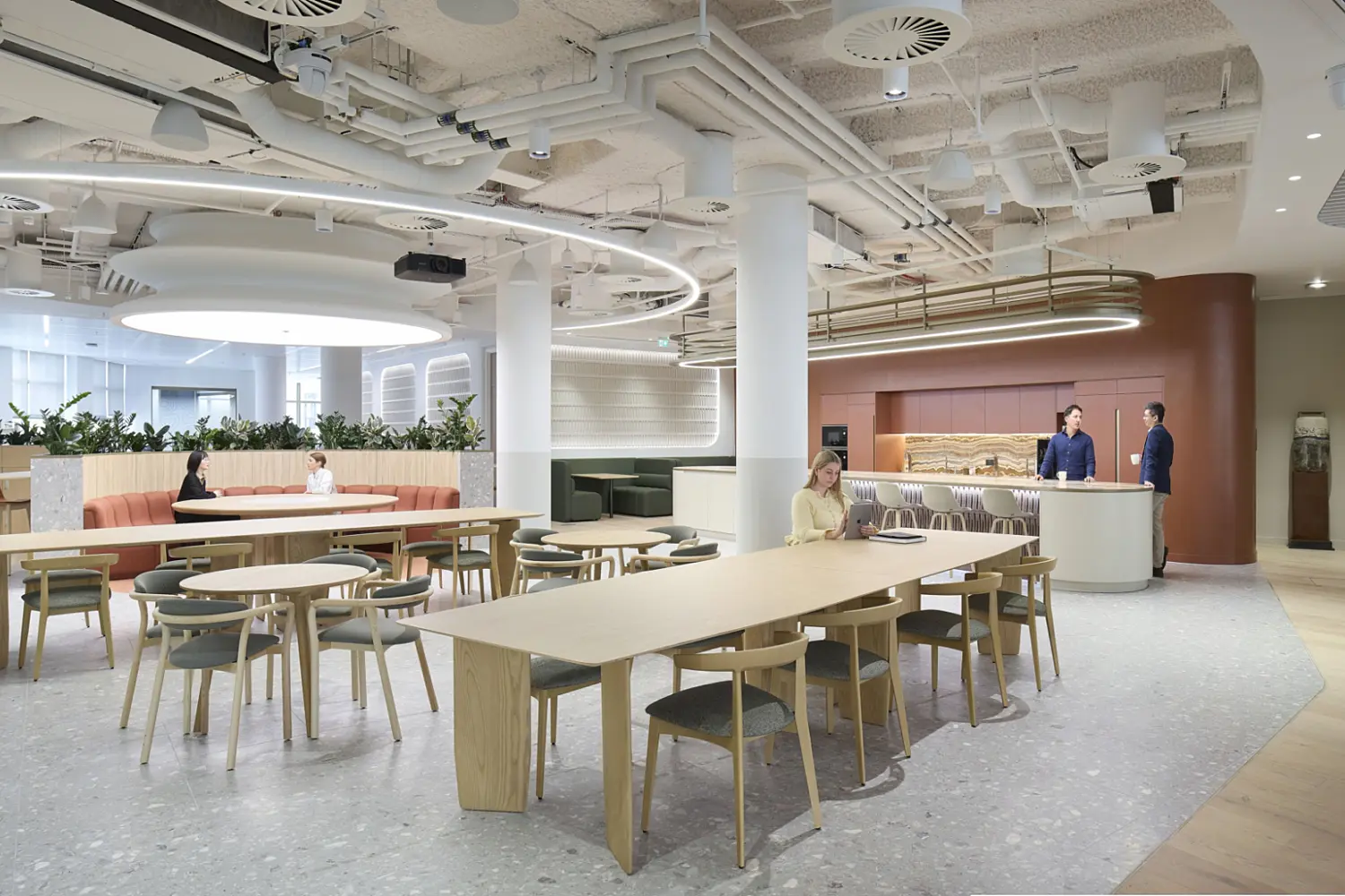 A spacious workplace dining and social area with long communal tables, round café tables and built‑in seating arranged around planting. Staff are seated, working and talking near the open kitchen counter in the background. Soft lighting, exposed ceiling details and natural materials create a relaxed multifunctional setting that reflects modern office fit out and office design.
