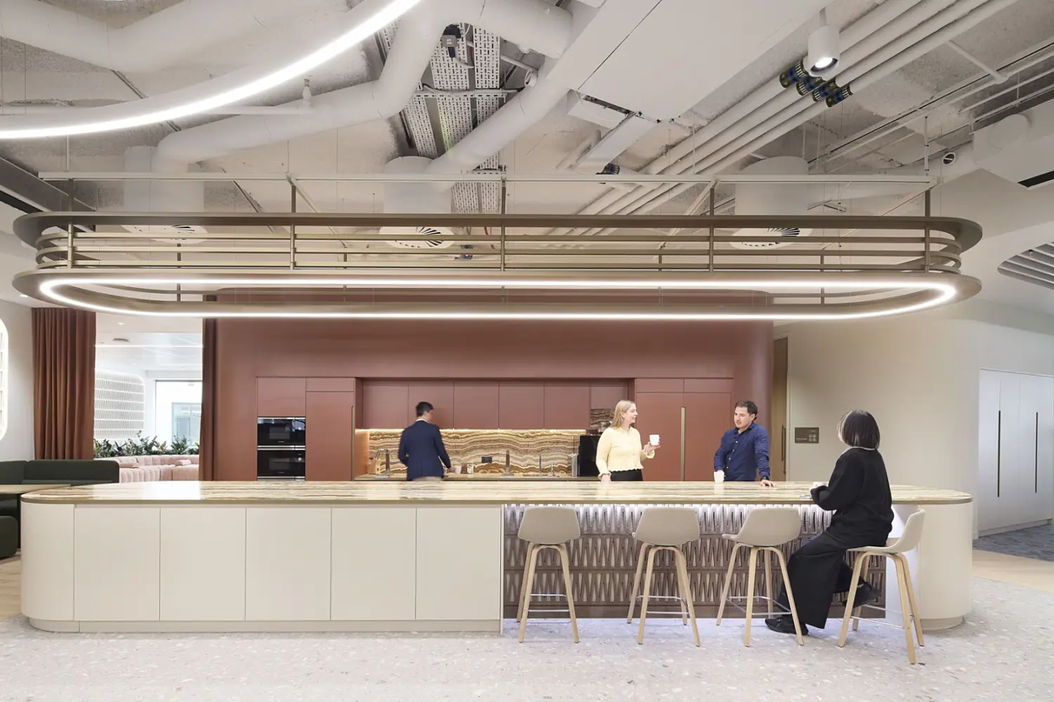 A modern workplace kitchen and social area with a long island counter, high stools and warm timber and stone finishes. Staff are gathered around the counter while another person prepares drinks behind it. Soft lighting, curved details and an open ceiling create a relaxed communal setting that reflects contemporary office fit out and office design.