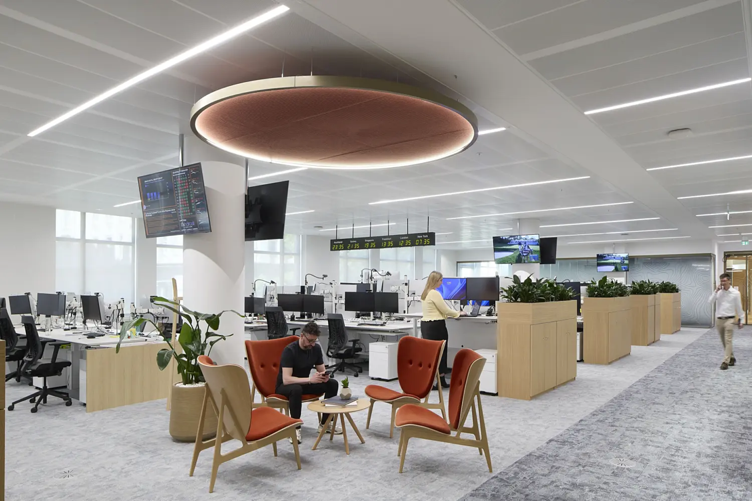An open plan workplace with rows of height adjustable desks, multiple screens and integrated planting between work zones. A breakout area with four orange lounge chairs and small tables sits beneath a large circular ceiling feature. Staff are working and walking through the space, which uses natural tones, soft lighting and organised layouts that reflect contemporary office fit out and office design.