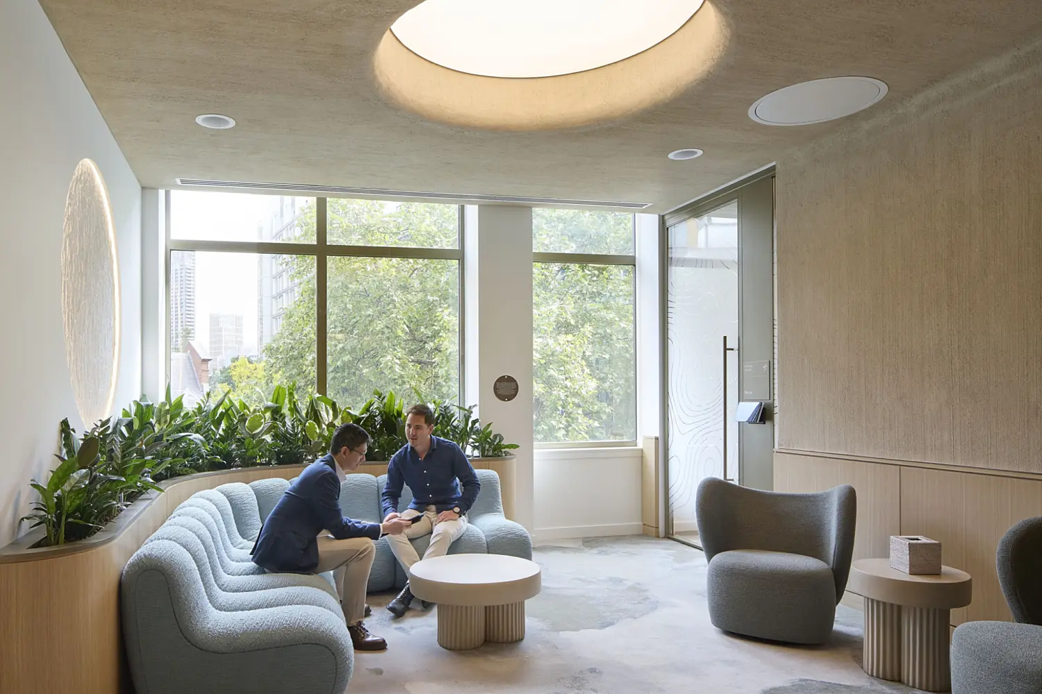 A bright lounge area with a sculpted ceiling light, large windows and a soft curved sofa where two people sit in discussion. The space includes contemporary armchairs, round tables and a line of planting along the wall. Natural textures, gentle lighting and a calm palette contribute to a modern workplace setting that reflects current office fit out and office design.