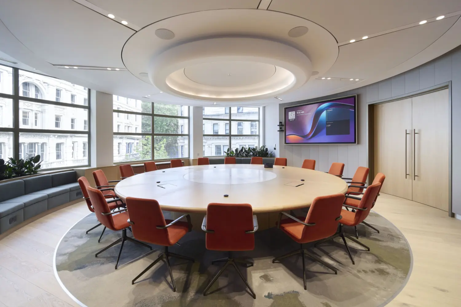 A circular meeting room with a large round table surrounded by orange swivel chairs, set within a bright space with full height windows. A sculptural ceiling feature sits above the table and a digital screen is mounted on the wall. Soft flooring, curved seating along the perimeter and planting help create a contemporary setting that reflects modern office fit out and office design.