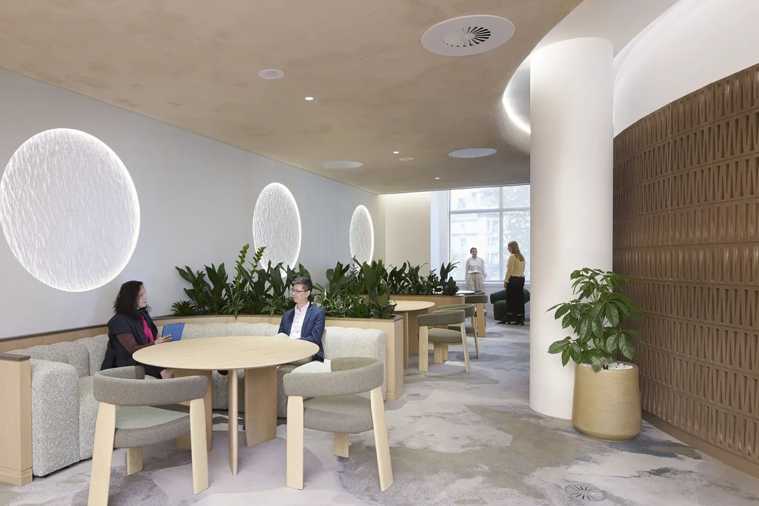 A calm workplace lounge with soft round tables, upholstered chairs and built‑in seating surrounded by abundant planting. Two people sit in conversation while others work further back in the space. Textured wall features, warm timber details and gentle lighting create a relaxed setting that reflects contemporary office fit out and office design.