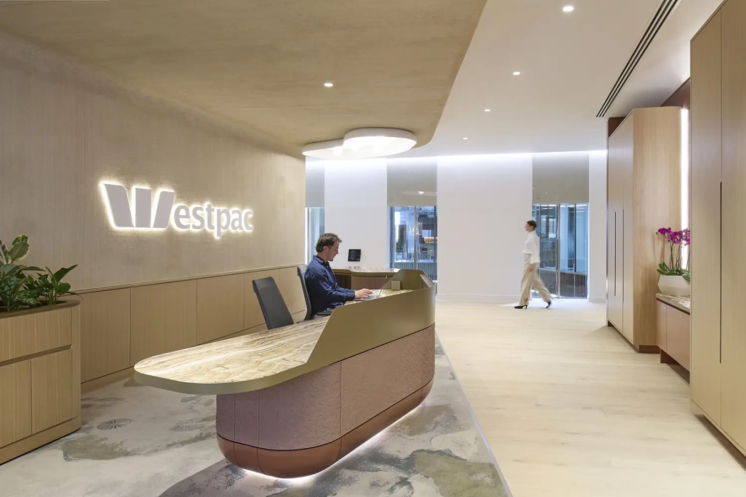 A contemporary office reception area created for Westpac, featuring a curved desk with a stone top and soft timber finishes. A receptionist is seated at the desk while a person walks through the bright open space beyond. Built-in storage, natural planting and subtle lighting contribute to a calm workplace environment that reflects modern office fit out and office design principles.