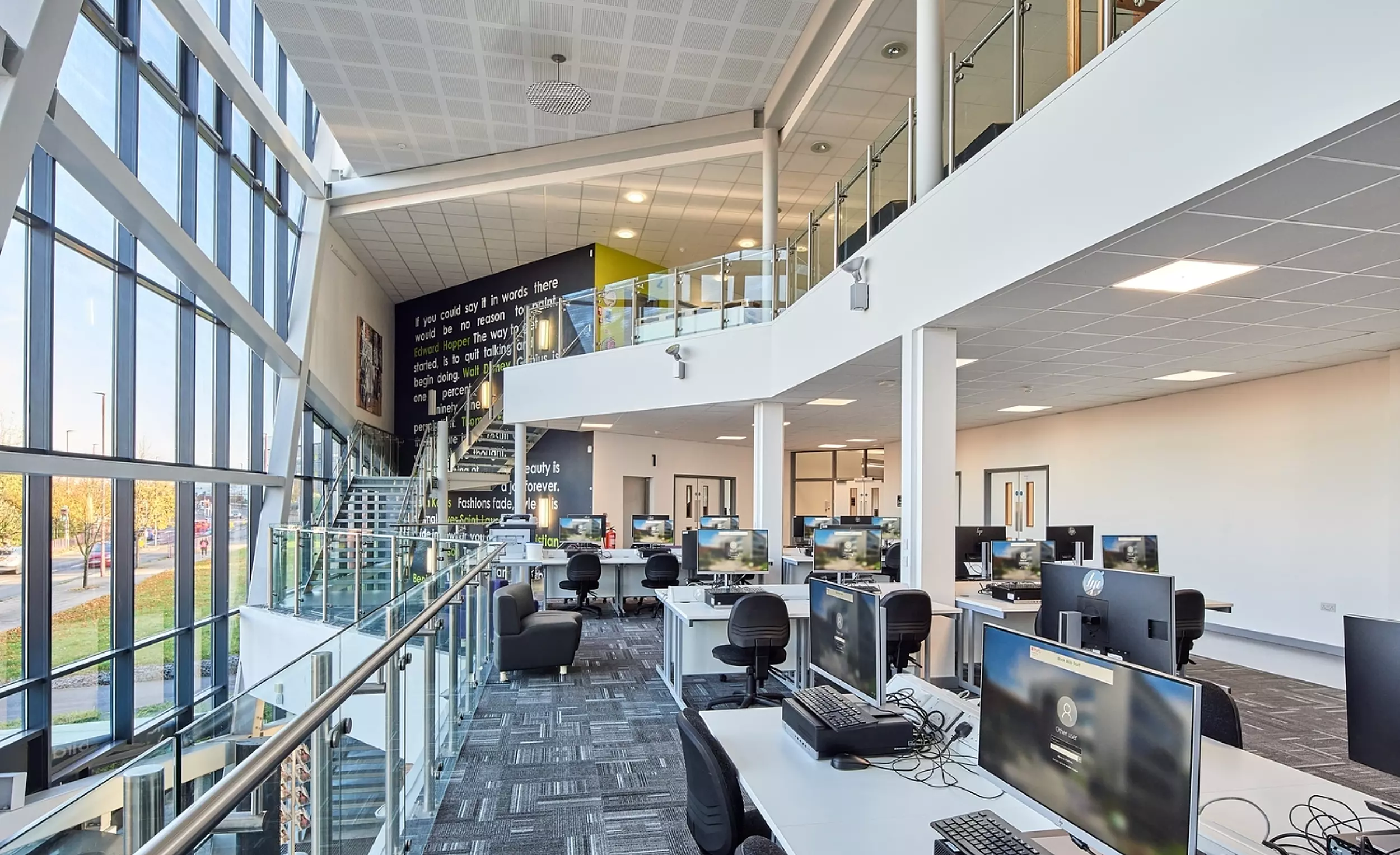 A modern, spacious university computer lab with large windows on the left letting in natural light. Multiple rows of desks with computers and chairs fill the room. A mezzanine level with glass railings provides additional workspaces. The ceiling features a grid pattern, and the floor is carpeted.